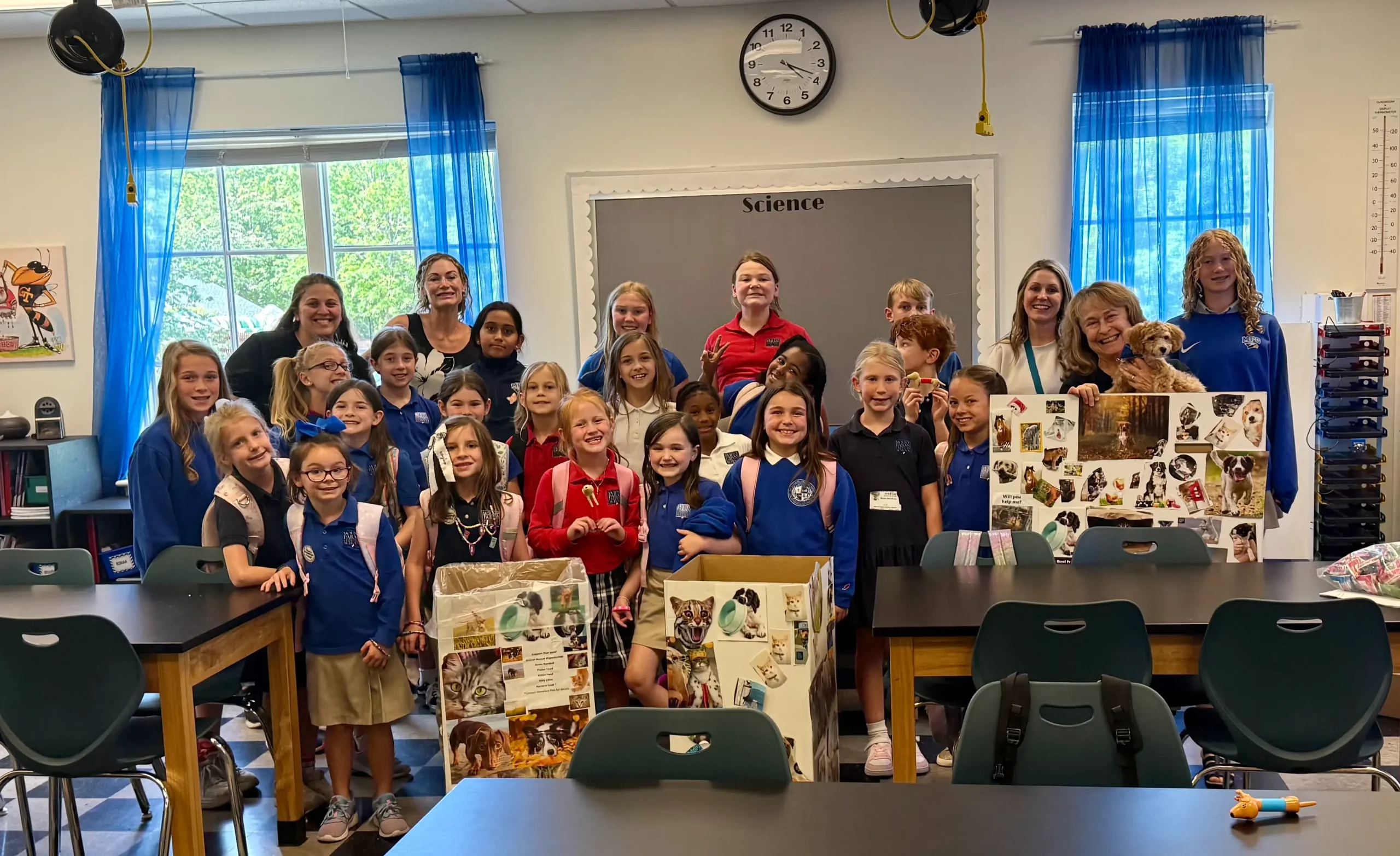 A large group of elementary school students display animal-welfare project posters in a classroom, alongside two humane educators and a small dog.