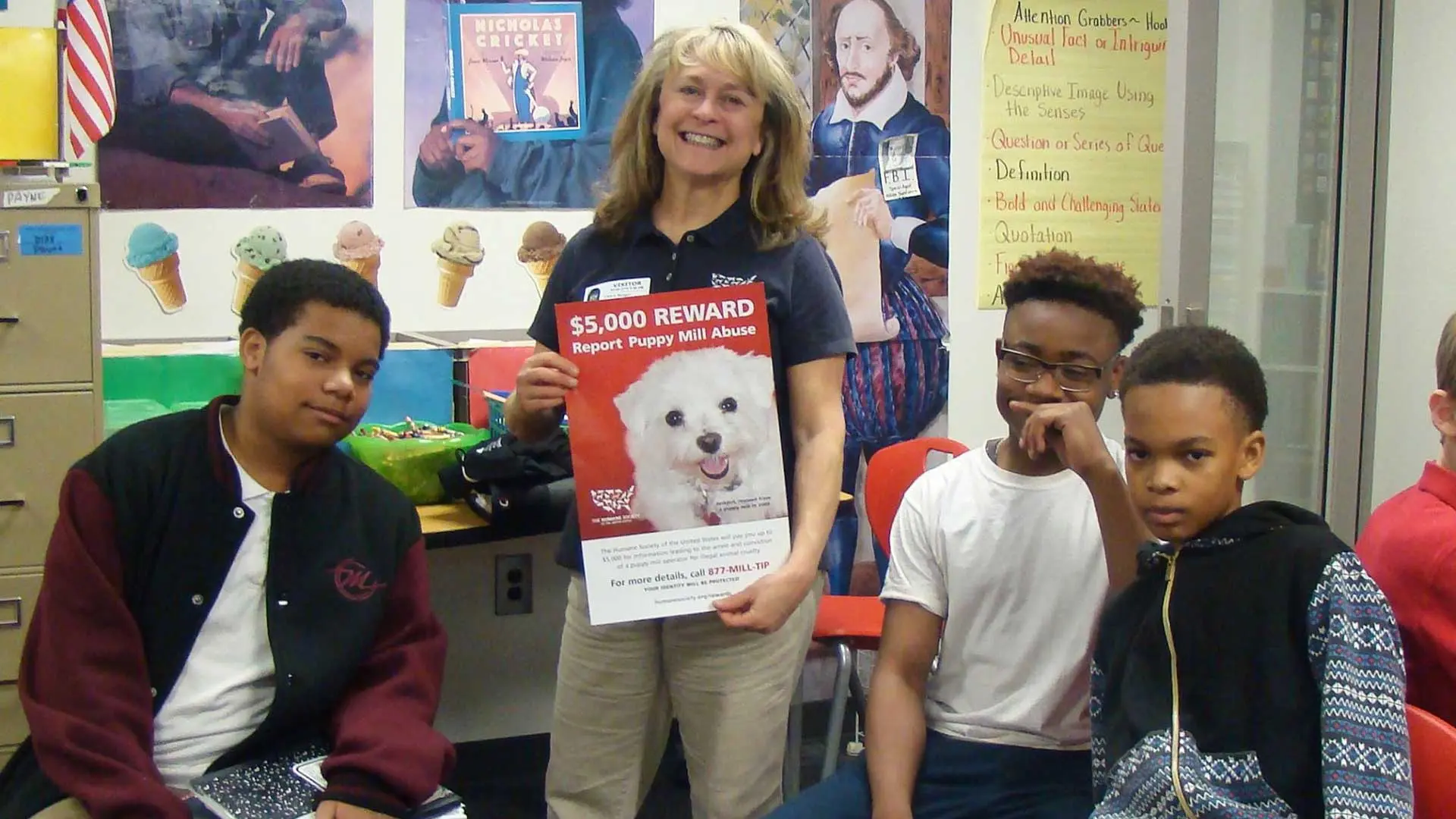 Humane educator holding a puppy-mill awareness poster while talking with middle school students in a classroom.